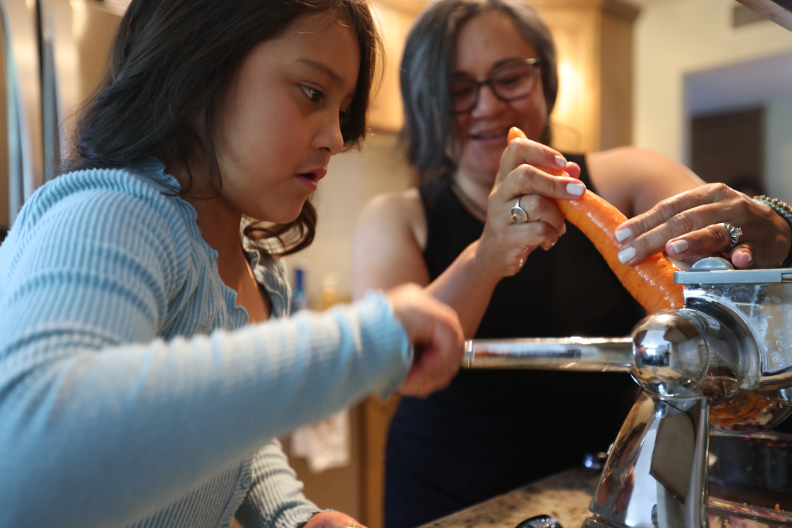 A grandmother helps her granddaughter grate a carrot.